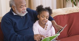 Young granddaughter sits on the lap of her grandfather as he reads a book to her