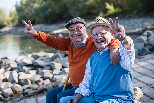 Two senior men sitting on the dock near the river while smiling and waving at the camera.