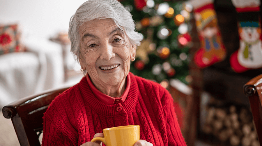 Older woman wearing a red sweater smiling at the camera while holding a yellow mug of tea.