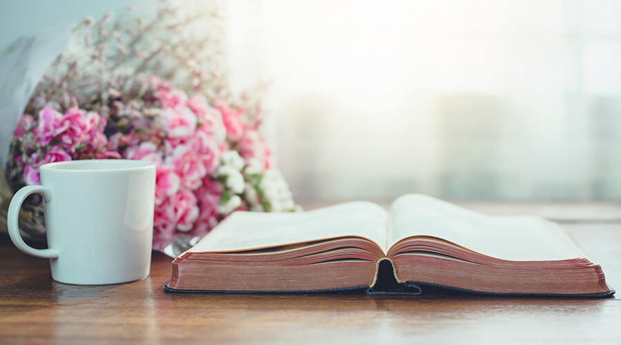 Light filtering in through window onto a table with a bible, coffee mug and flowers.