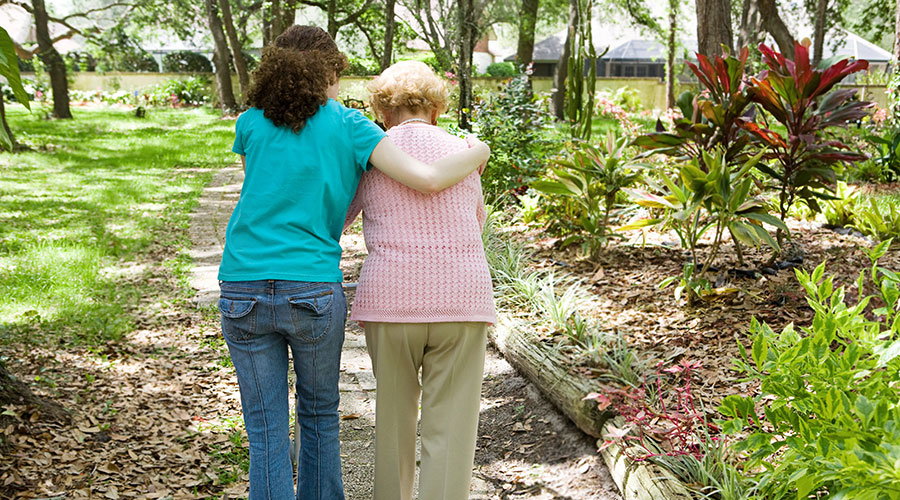 Senior woman being assisted walking by a woman outside on a trail.